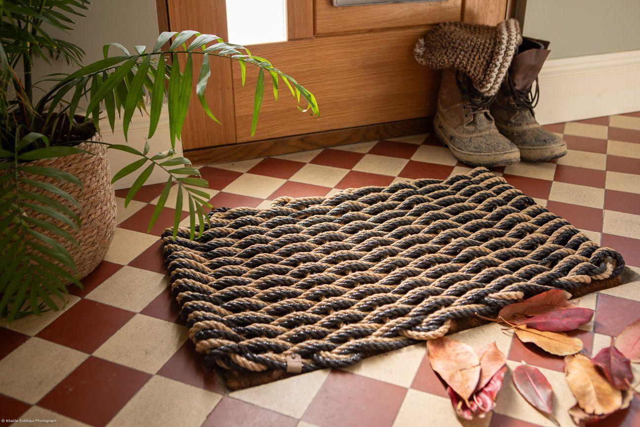 A handwoven rope doormat with a geometric pattern placed on a tiled floor, featuring a door in the background along with boots and a plant.